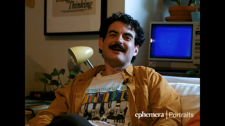 A man in his 20s with a mustache and wavy hair smiling at the camera.